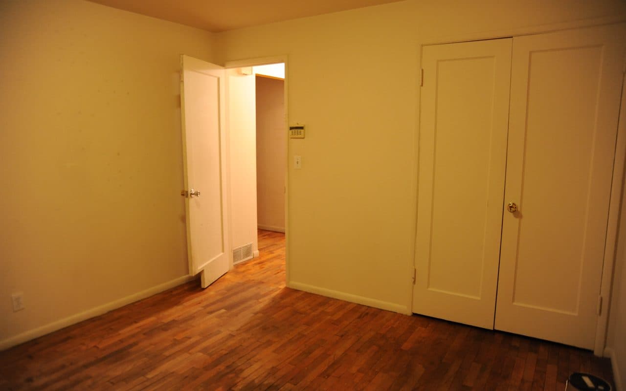 Empty bedroom at night with an open door onto a hallway, a closet with two doors, an alarm panel and a light switch on the wall.