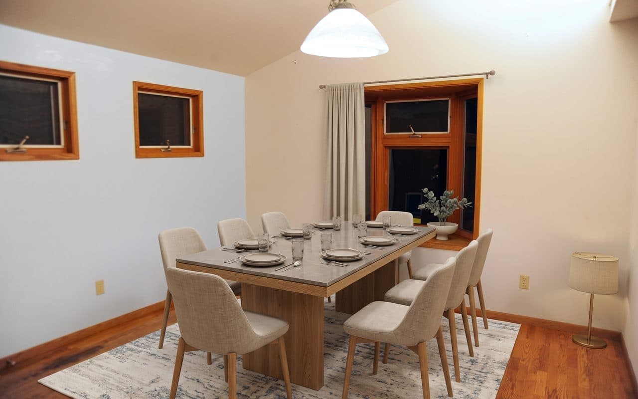 The same dining room with a long table set with plates, a floor lamp, and a pale curtain on a rod hung across the left side of the bay window.