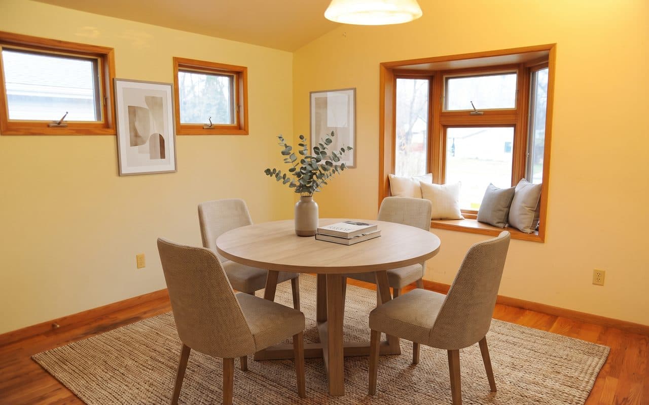 A dining room in daylight with a round table and four chairs, a larger bay window with a cushioned seat, and bigger windows on the left wall.