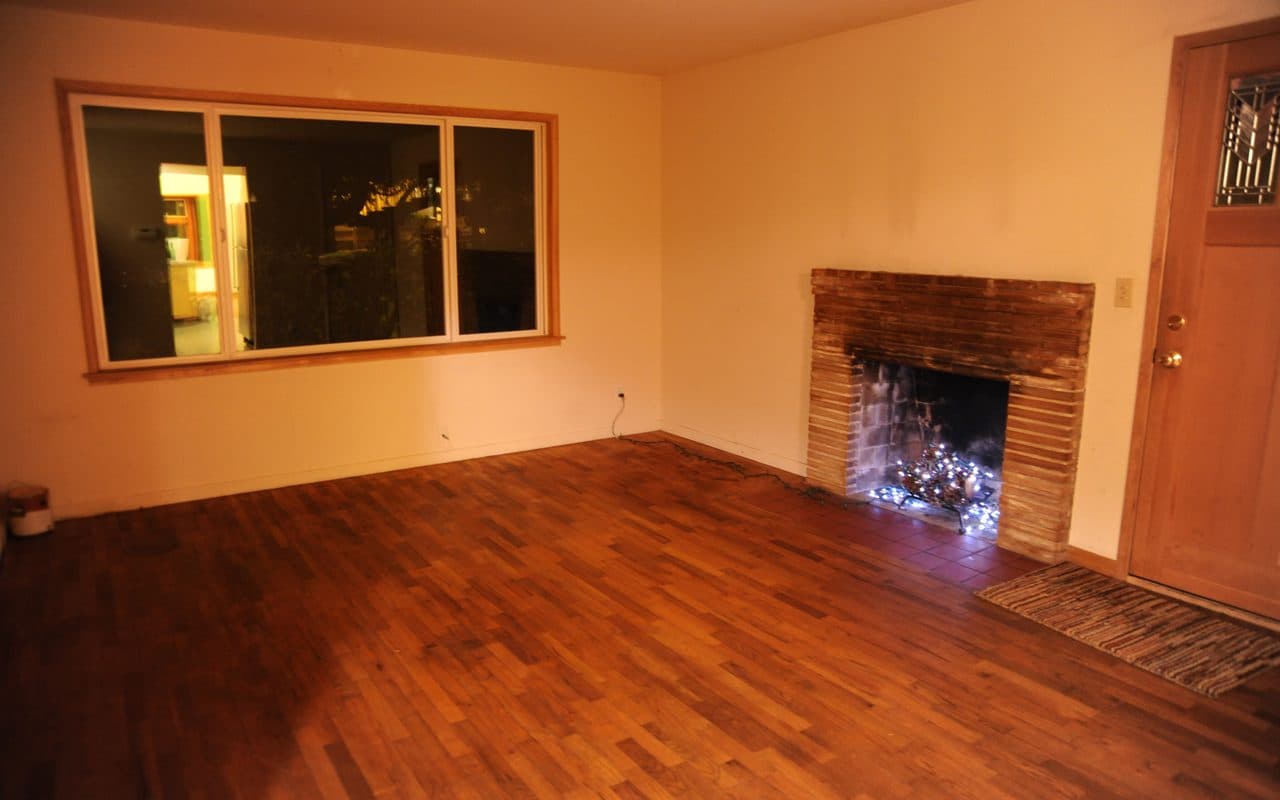 Empty living room at night with a wood floor, a large dark window, a brick fireplace holding a string of lights, and a door on the right.
