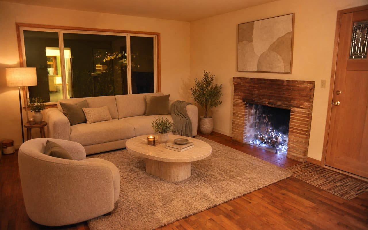 The same living room with a sofa under the window, a round stone coffee table, a curved armchair, a floor lamp and art above the fireplace.