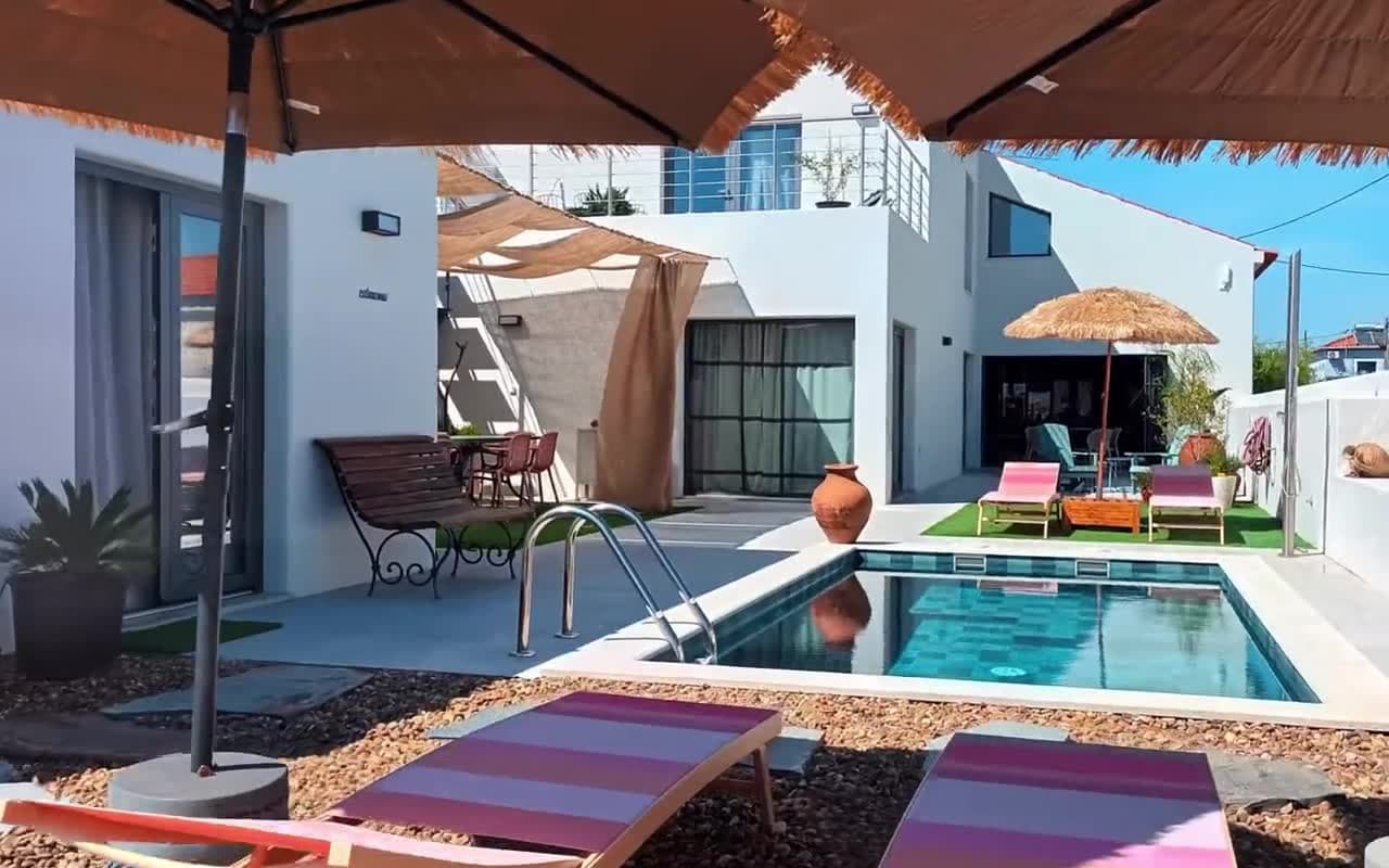 Frame from an AI walkthrough clip: a white house around a small pool, with straw umbrellas, pink sun loungers on gravel and a shaded seating area.