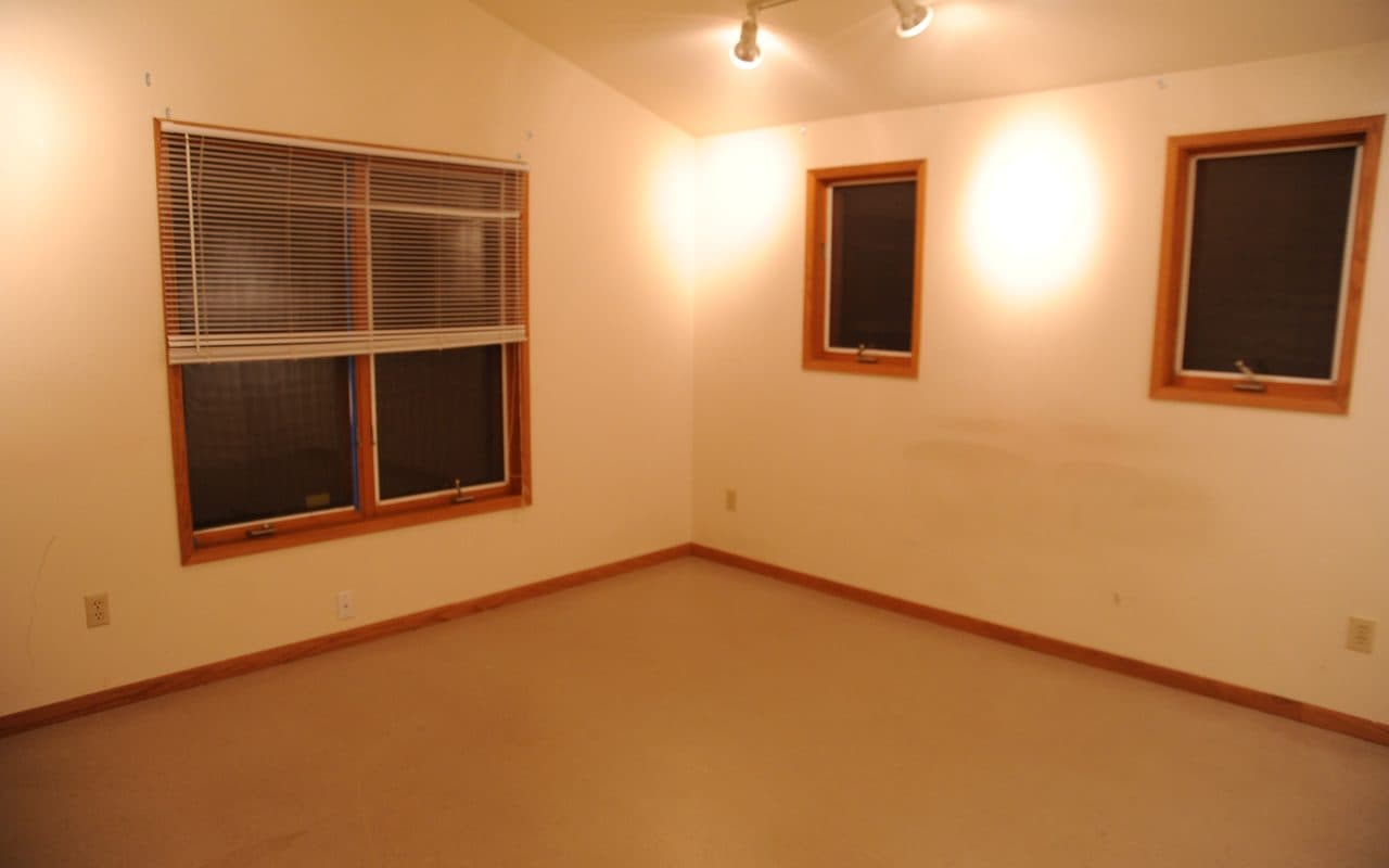 Empty carpeted room at night with a sloped ceiling, track lights, a window with blinds on the left and two small windows on the right.