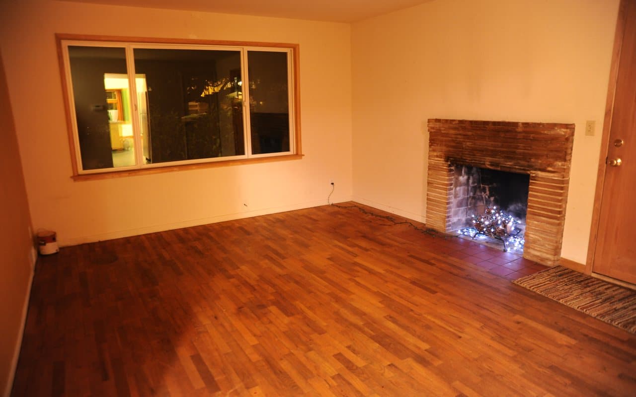 Empty living room at night with a wood floor, a large dark window, a brick fireplace holding a string of lights, and a door on the right.