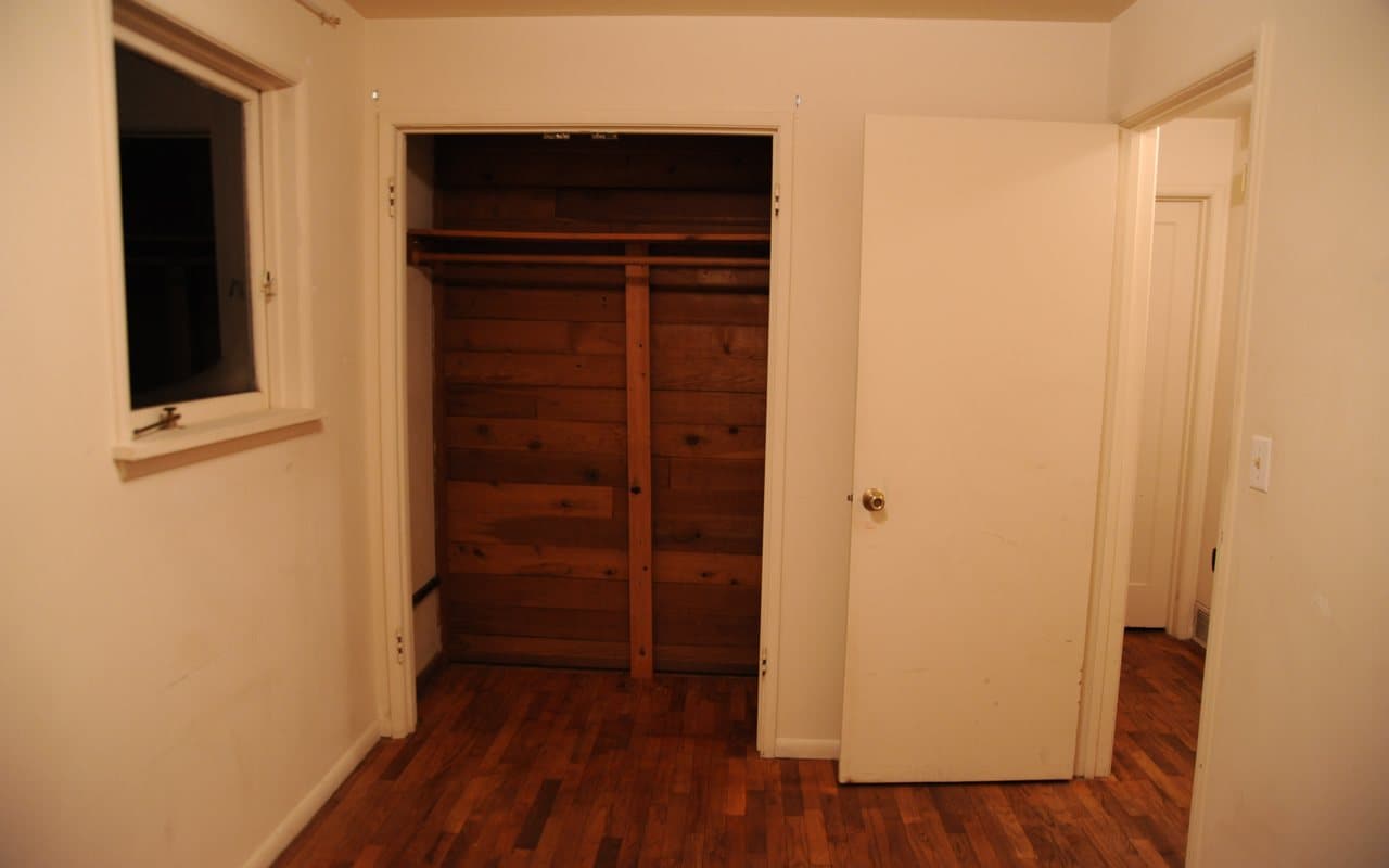 Small empty bedroom at night with a window on the left, an open wood-lined closet, a white door standing open and a hallway beyond.