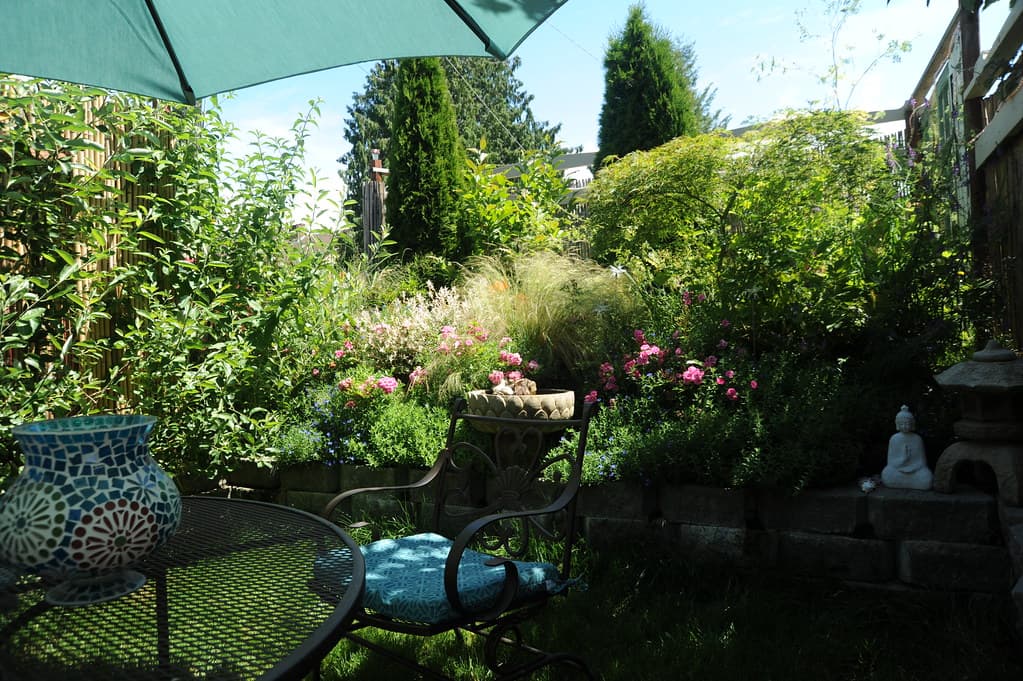 Garden patio with a blue umbrella over a round table and chairs, surrounded by shrubs, flowering plants and tall trees.