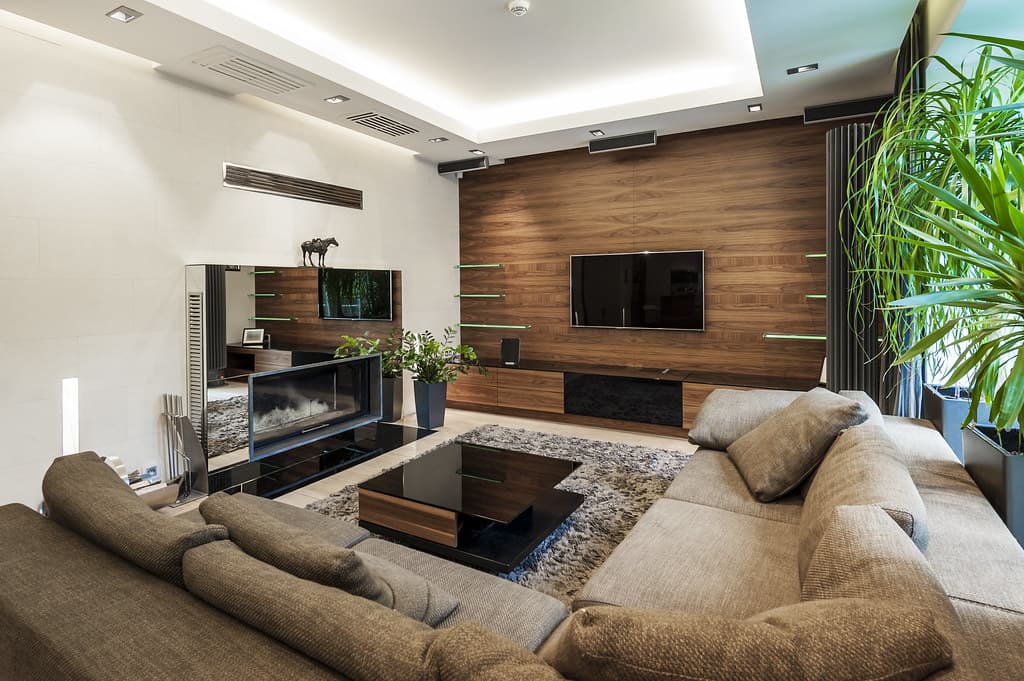 Furnished living room with a taupe sectional sofa, a black glass coffee table on a shaggy rug, a walnut-paneled TV wall and a glass-fronted fireplace.