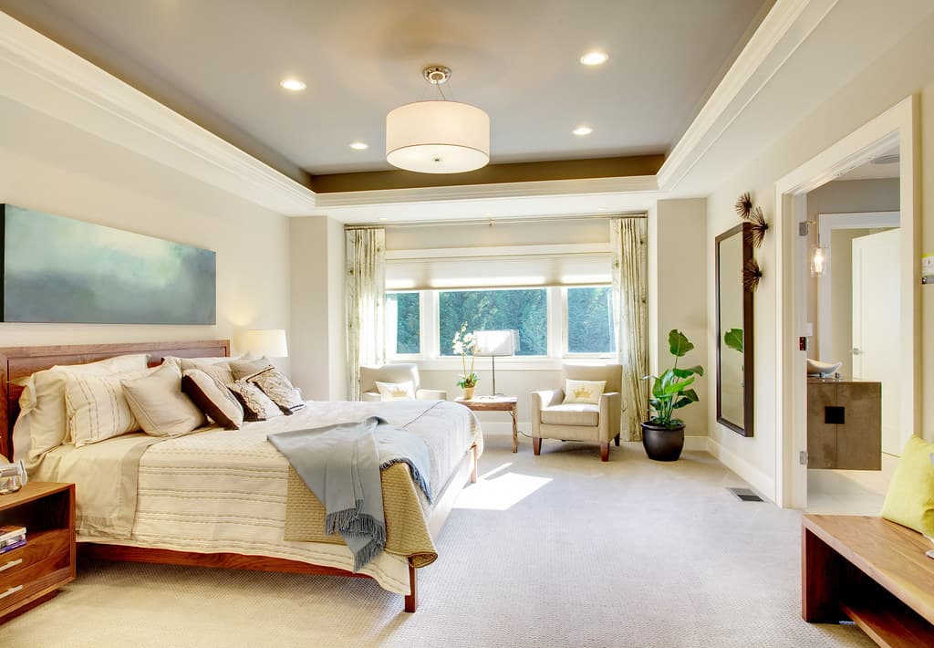 Bright bedroom with a large made-up bed under a wide painting, a lamp on the bedside table, two armchairs by the window and a plant.