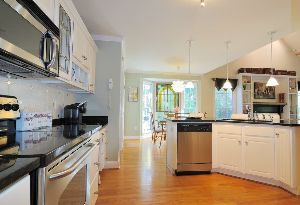 Kitchen with white cabinets, black counters, stainless steel appliances, pendant lights over a peninsula and a honey-colored wood floor.