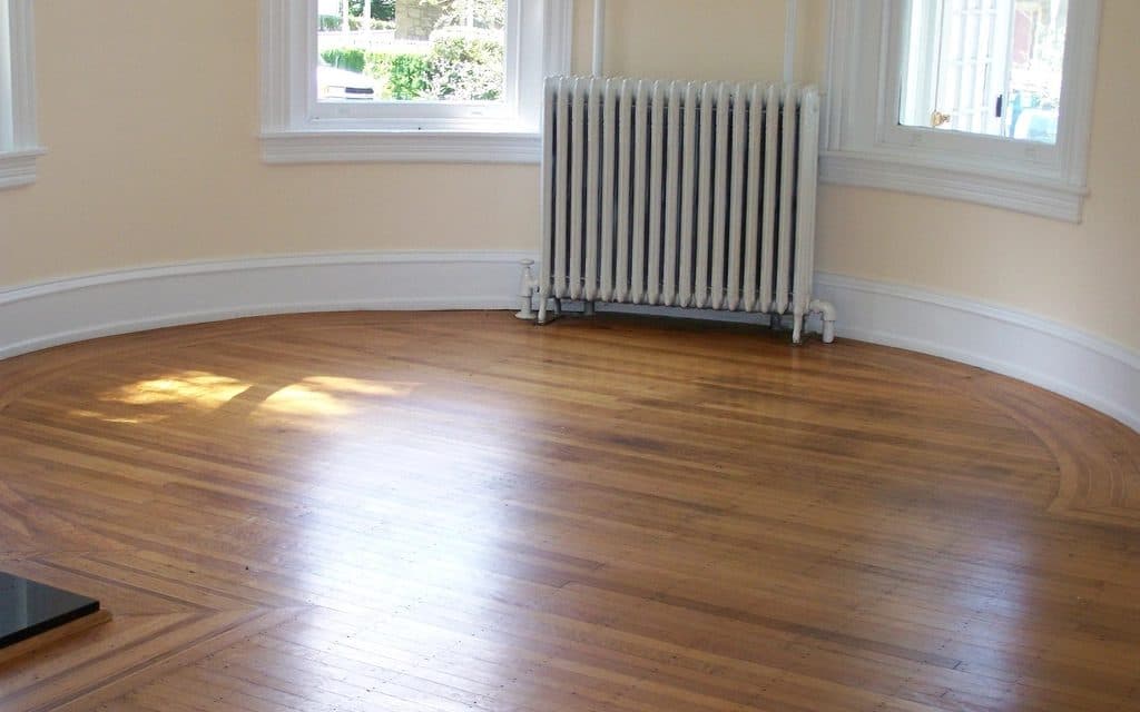 Empty living room with a curved bay wall, two windows, a white radiator and patches of direct sunlight on the wooden floor.