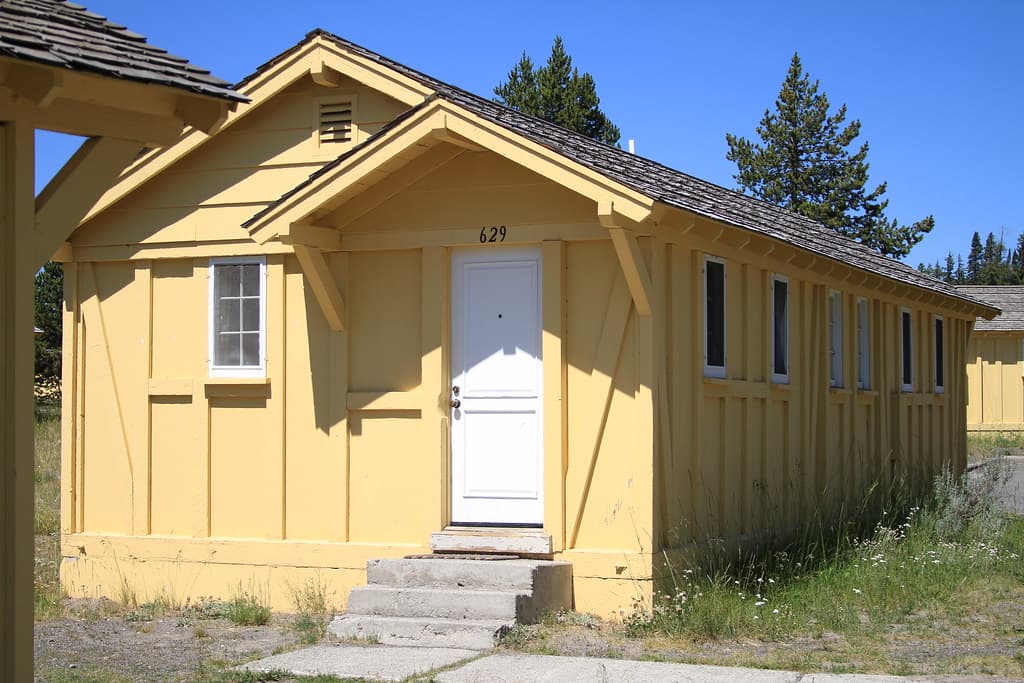 Front of a yellow single-storey wooden cabin with white window trim, a white front door and a short run of steps, under a blue sky.